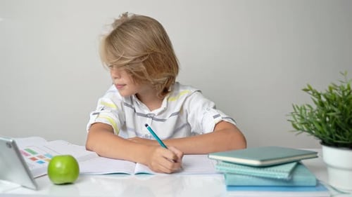 Middle School Smiling Student Boy Sitting at Desk Studying Writing Book Homework and Tablet at Class