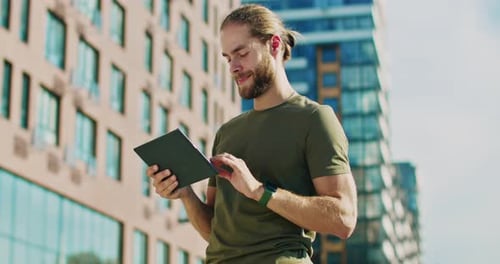 Kaukasischer lächelnder junger gutaussehender Mann in khakifarbenem T-Shirt steht draußen und benutzt ein Tablet-Gerät
