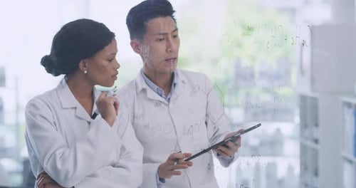 Two scientists using a digital tablet while brainstorming equations and formulas on a glass wall