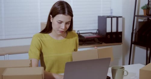 Asian woman checking laptop screen while holding parcel box in home office, preparing online orders