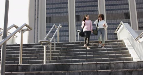Diverse female friends descending staircase at urban plaza gesturing and talking with duffel bags