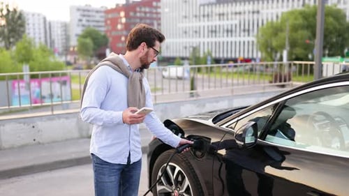 Man Using Phone While Charging Electric Car Outdoors