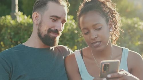 Close up, happy smiling interracial couple watching video on mobile phone while sitting on bench