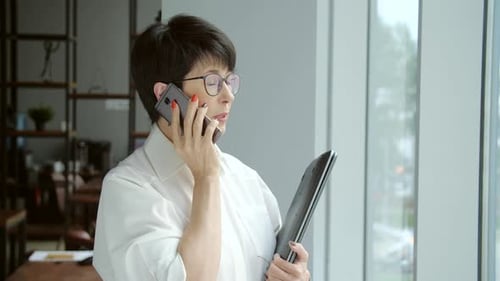 Office Work Concept A Mature Businesswoman Stands By the Window and Talks on the Phone