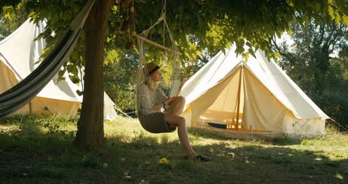 Woman Reads Book in Hanging Chair Near Tents