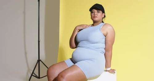 Entering frame woman sitting on cube adjusting cap settling for studio shoot against split backdrop