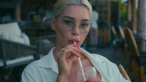 Blonde Girl in White Shirt is Working with Laptop in Cafe Looking at Camera and Drinks Shake