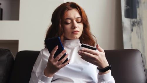 Smiling Young Woman Customer Holding Credit Card and Smartphone Sitting on Couch at Home Happy