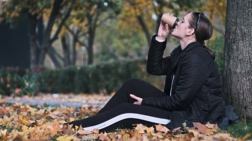 Young Woman Drinks Coffee on Lawn in Autumn Park Among Fallen Yellow Leaves