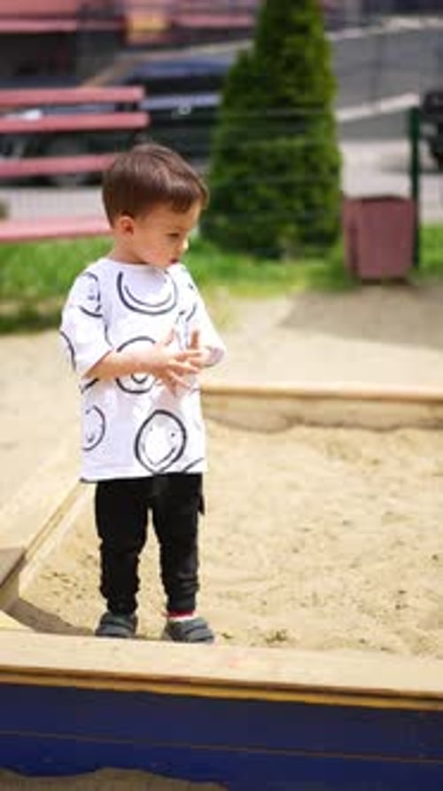 Little toddler sitting in the sand box. Baby boy stands up pouring some sand from hands.