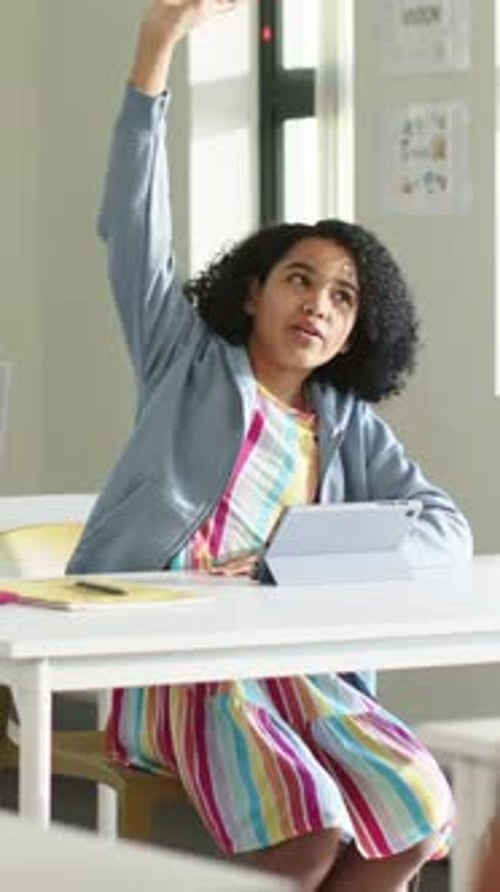 Vertical video: Teacher cueing student in striped dress raising hand at school with tablet to speak