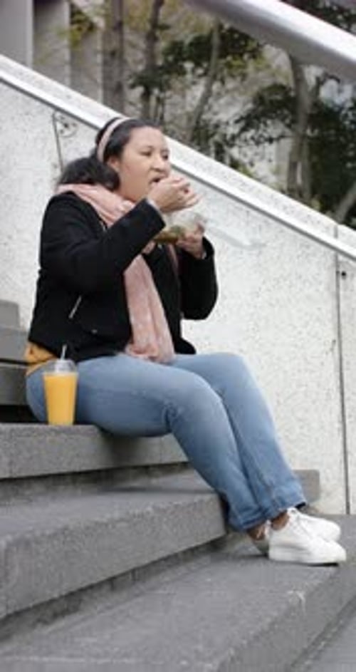 Vertical video: Mature woman peeling salad container, sampling lunch and glancing passerby on steps