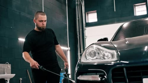A male car wash employee applies car wash detergent to a black luxury car using spray gun in the car