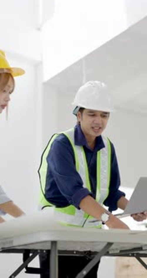 Asian female engineer holds laptop and looks upward beside colleague, discussing ceiling work,