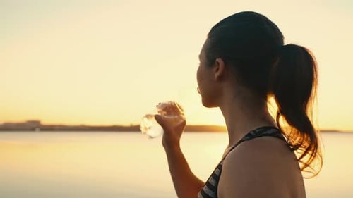 Portrait of an Athlete Girl Drinking Water From a Bottle at Sunset By the Lake