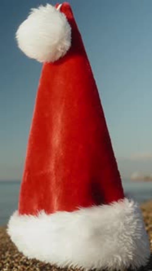 Vertical video. The red Santa hat is placed on the sandy beach by the sea on an early sunny morning,