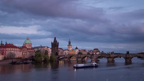Day to night sunset timelapse along the Charles Bridge on the Vltava river with boats on a cloudy ev