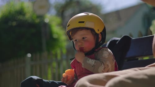 Toddler in yellow helmet sitting on bench outdoors, tasting tangerine segment then pausing to