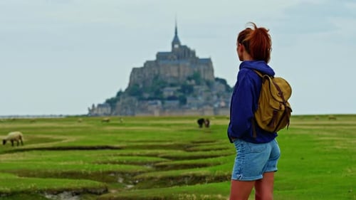 A Female with a Backpack Visits the Mont Saint Michel Castle While Walking Through Green Meadows
