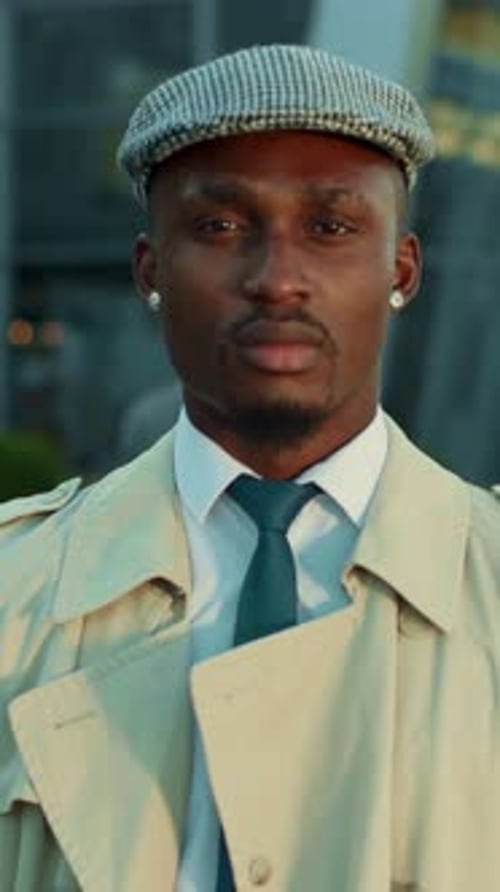 A Stylish Man Dressed in a Tan Trench Coat and Wearing a Cap Stands in an Urban Location