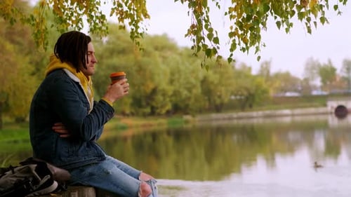 Young Handsome Man with Dreadlocks in Warm Clothes Drinking Coffee Near Lake in Golden Autumn
