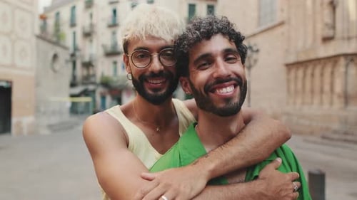 Gay couple stands hugging and looking at camera on street of old city
