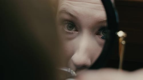 Young blonde caucasian girl applying an eye mascara in the mirror, close up