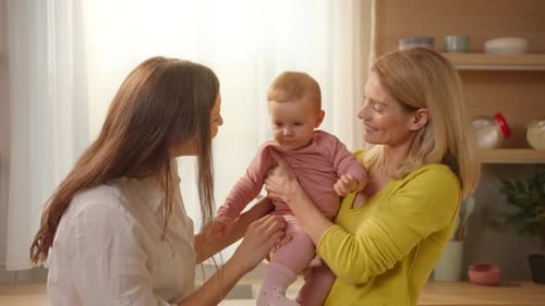 Young Cheerful Caucasian Woman in Love Holding Little Baby in Pink Clothes Indoors