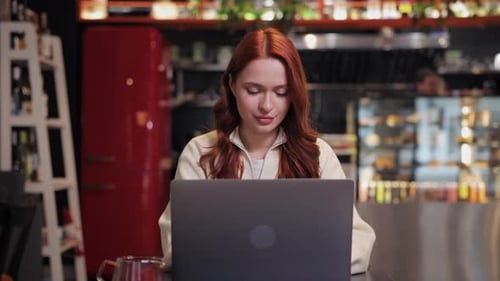 Concentrated Redhaired Woman Sitting in Modern Cafe Working on Laptop Typing Carefully Focusing on