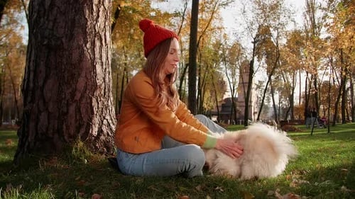 Cheerful Girl in a Hat Hugging White Pomeranian Spitz Dog in the Park