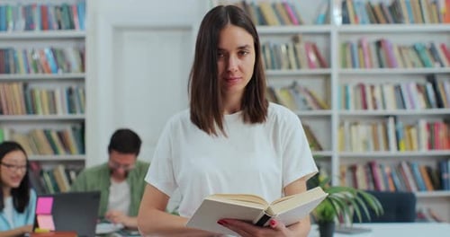 Portrait of Beautiful Young Girl Holding Book in Library Bookshelf Background Looking at the Camera