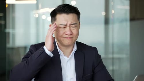 Asian businessman suffering from headache while sitting at workplace in modern business office.