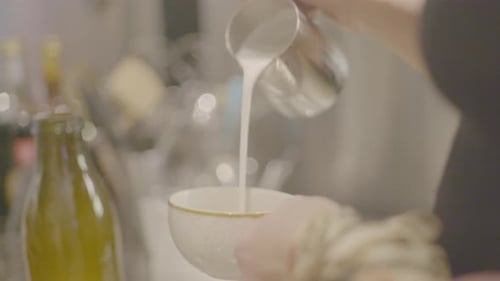 Barista gießt langsam eine Tasse Milch in eine Tasse Kaffee mit mehr Dosen und einer Kaffeemaschine in der