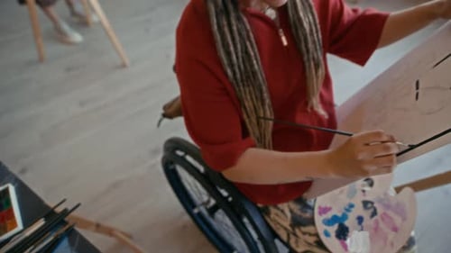 Young Female Artist in Wheelchair Holding Canvas and Painting in Studio