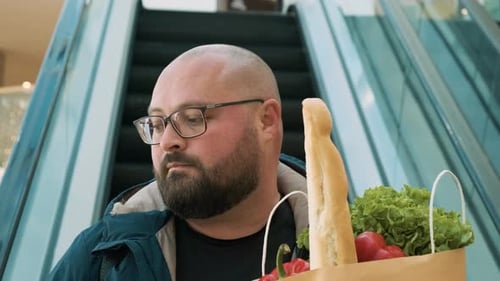 Homme en surpoids sur un escalator avec des aliments diététiques dans un emballage écologique dans un centre commercial Sustainable Lyfestyle