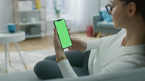 Young Woman at Home Uses Green Mock-up Screen Smartphone. She's Sitting On a Couch in His Cozy Livi
