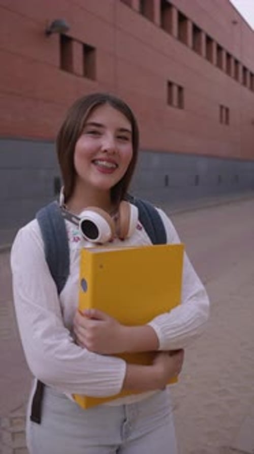 Female student carrying yellow binder and backpack standing confidently near school building, radiat