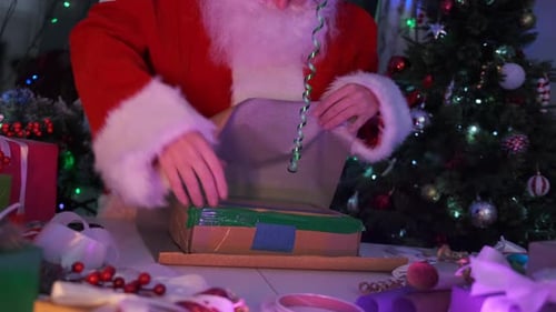 Close-up of gift box wrapping Santa hands in paper at table in workshop, preparing to go congratulat