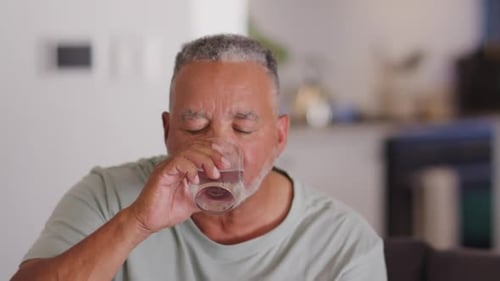Älterer afrikanischer Mann hat Durst, hebt klares Glas, trinkt langsam auf dem Sofa zu Hause und lächelt