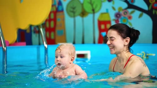 Adorable chubby baby boy making splashes in the pool. Woman is holding a child teaching him to swim.
