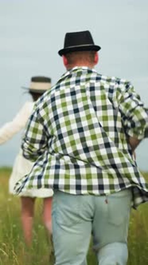 Man in Hat Chasing Woman in White Dress Through Grassy Field on Cloudy Day
