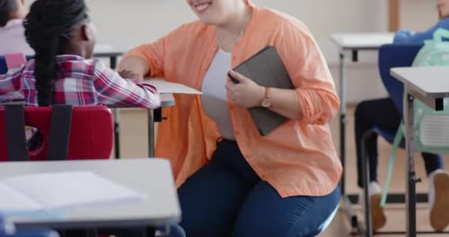 In school, female teacher holding tablet smiling and talking with student in classroom