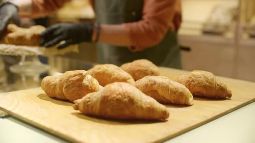 Gloved Hands of Female Baker Putting Croissants on Cake Stand on Shop Counter