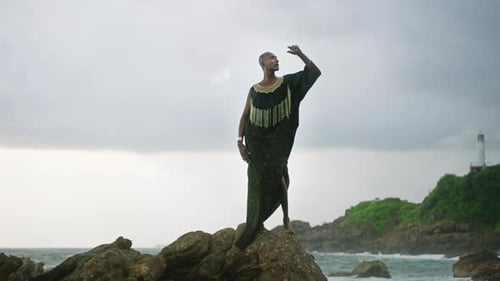 LGBTQIA Black Person in Boutique Dress Jewelry Stands on Rock on Stunning Ocean Coastline Skyline