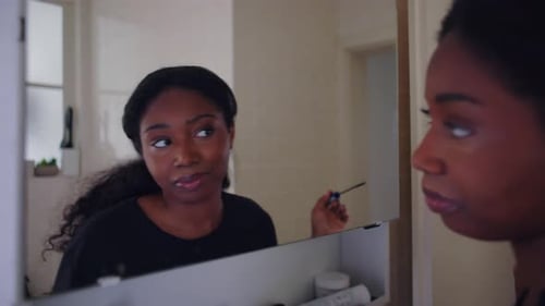 African American woman applying mascara to eyelashes while looking at her own reflection in