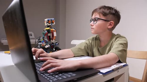 Young Boy with Glasses Focused at a Laptop Keyboard Coding and Assembling a Small Robot on His Desk