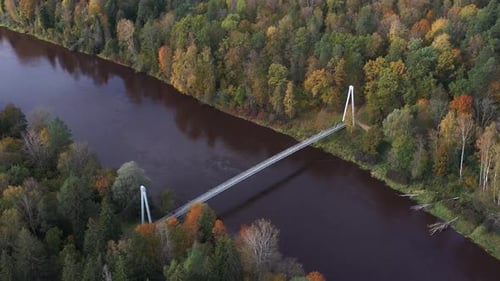 A drone glides over a bridge spanning a winding autumn river, revealing vibrant fall trees in a colo