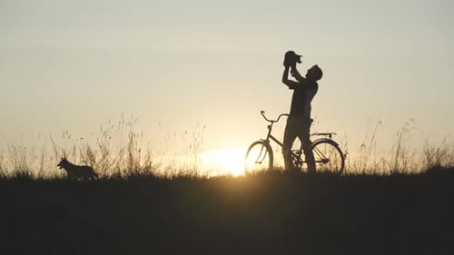 A silhueta de um homem com uma bicicleta levanta seu amado cachorro até o topo ao pôr do sol