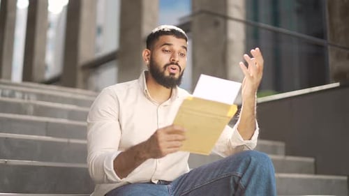 Disappointed adult man reading letter with bad news while sitting on the street on the steps