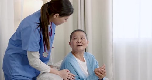 Young asian nurse kneeling beside senior cancer survivor during homecare rehabilitation, providing